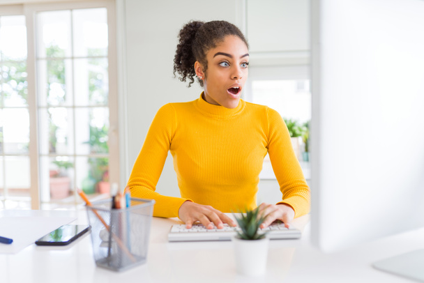 Fiatal afroamerikai lány dolgozik számítógéppel félek a sokk egy meglepetés arc, fél és izgatott félelem kifejezés - Fotó, kép