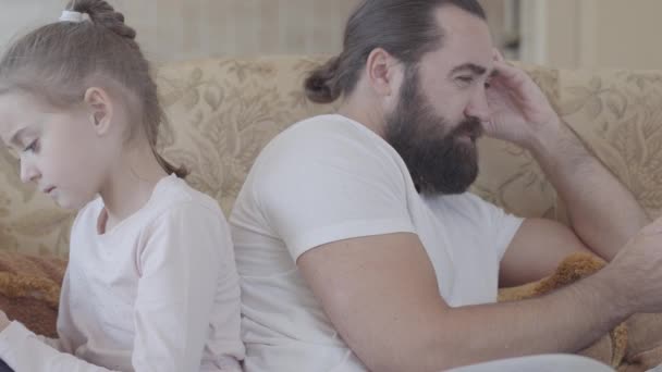Close up portrait of cute daughter and her father sitting on the sofa on living room with cell phones and chilling in cozy day. Father and daughter relationship. - Materiał filmowy, wideo