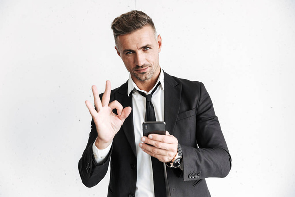 Business man posing isolated over white wall background using mobile phone showing okay gesture. - Foto, imagen