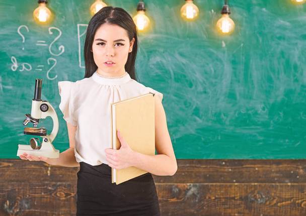 Biology concept. Teacher of biology holds book and microscope. Lady in formal wear on calm face in classroom. Lady scientist holds book and microscope, chalkboard on background, copy space - Фото, изображение