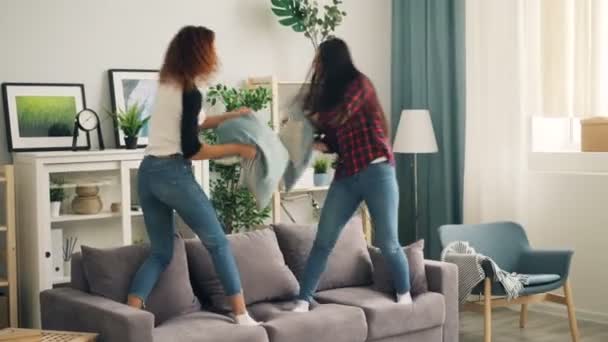 Playful young women African American and Asian are fighting with pillows standing on sofa and laughing. Girls are wearing casual clothing shirts and jeans. - Кадри, відео