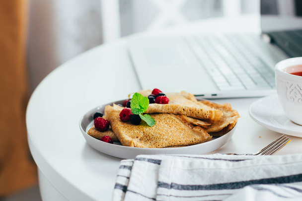Delicious Breakfast. White desktop table with laptop and fresh crepes pancakes, cup of tea. Freelancer concept. Healthy breakfast food. - Fotografie, Obrázek