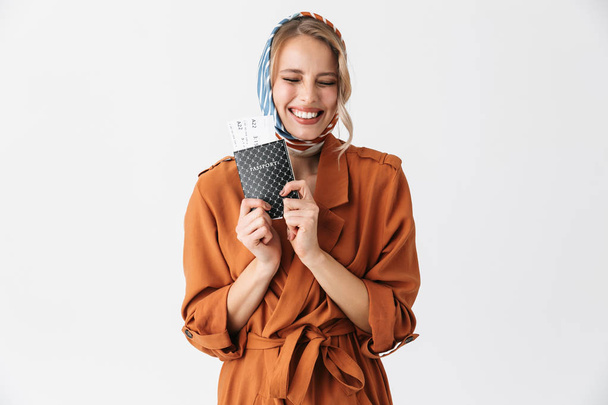 Beautiful excited young blonde pretty woman wearing silk scarf posing isolated over white wall background holding passport with tickets. - Fotó, kép