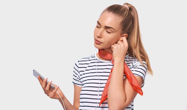 Pretty European young woman with ponytail hairstyle, wearing striped t-shirt, stylish red scarf on neck, relaxing with closed eyes listening to her favourite songs via white earphones using smartphone - Foto, Imagen