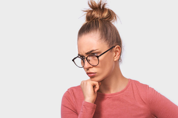 Thoughtful blonde beautiful young woman wears eyeglasses and casual pink blouse, looks seriously away to someone, poses against white studio background. People emotions concept - Foto, immagini