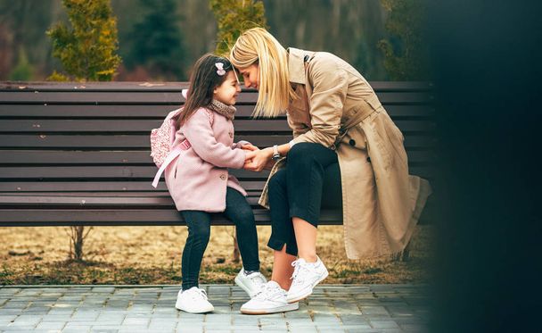 Candid horizontal image of beautiful mother and her girl kid smiling, sitting on a bench outdoors. Loving joyful young woman and her happy daughter spending time together in the city park. Mothers Day - Photo, image