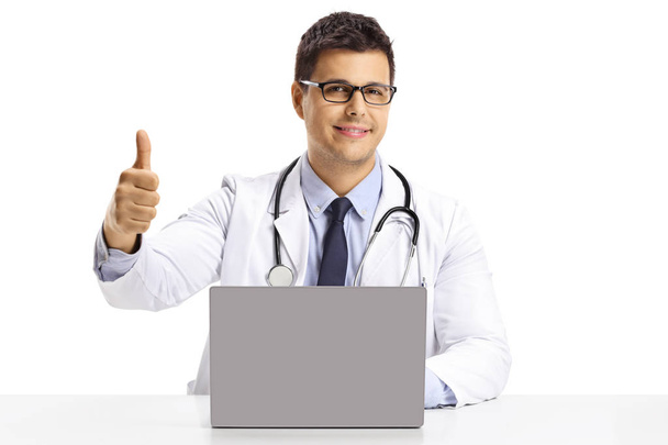 Young male doctor sitting with a laptop and showing thumbs up isolated on white background - Foto, Bild