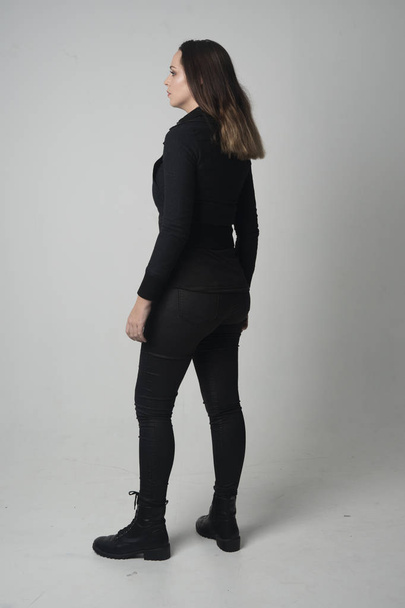 full length portrait of a brunette girl wearing  modern black jacket and pants, standing pose with back to the camera on grey studio background. - Foto, afbeelding