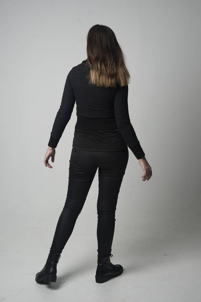 портрет брюнетки в современной черной куртке и брюках, стоящей в позе спиной к камере на сером фоне студии
. - Фото, изображение