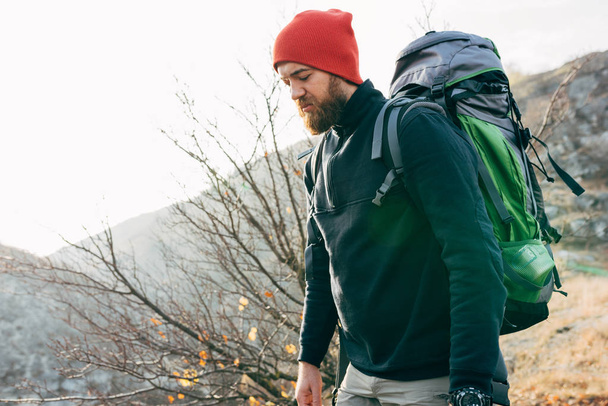 Side view outdoors image of bearded young man hiking in mountains with big travel backpack. Traveler male in red hat trekking during his journey. Travel, people, sport and healthy lifestyle concept - Фото, зображення