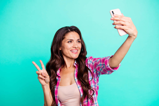 Close up photo beautiful she her lady hold hand arm smart phone new model make take selfies show v-sign say hi wearing casual plaid checkered pink shirt outfit isolated teal bright background - 写真・画像