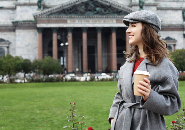 Smiling stylish young woman drinking coffee while walking on a city street - Foto, Bild