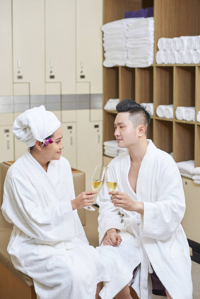 Fiatal ázsiai pár ül a fürdőköpeny és pirítás pohár pezsgő öltözőben wellness-központban - Fotó, kép
