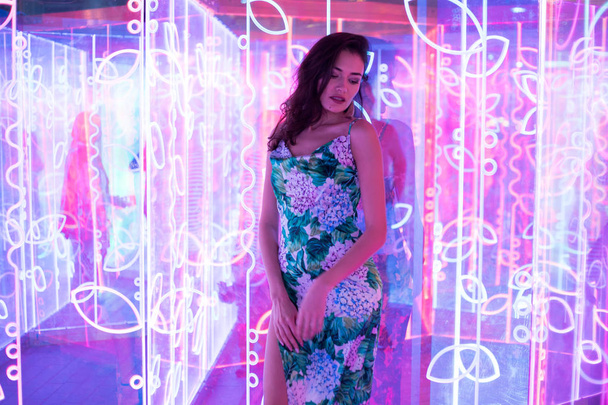Beautiful woman in a dress with a floral print stands on the background of neon lights. - Photo, image