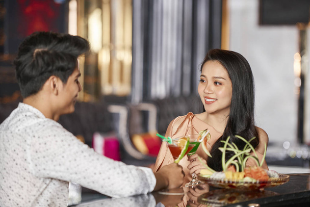 Happy Asian couple celebrating Valentines Day with cocktails while sitting at bar counter at restaurant - Photo, Image