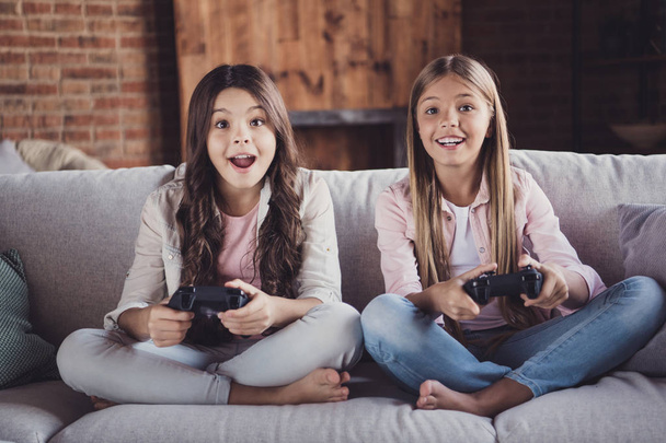 Portrait of two nice crazy lovely attractive charming funny cheerful cheery girls sitting on divan playing video game device crossed legs in house loft industrial interior style - Foto, imagen