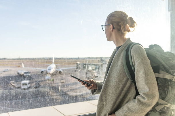 Satunnaisesti pukeutunut naispuolinen matkustaja lentokentällä, jolla on älypuhelin laite, katselee lentokentän portin ikkunoiden läpi lentokenttien kiitoradalla - Valokuva, kuva