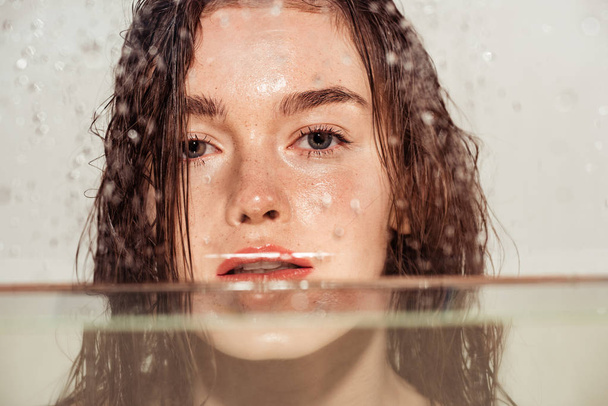 piękna młoda kobieta z korala usta patrząc na kamery przez szkło z kropli wody na szarym tle - Zdjęcie, obraz