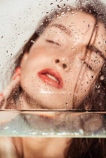 beautiful young woman with coral lips posing through glass with water drops isolated on grey - Photo, Image