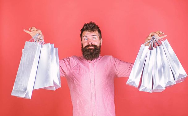 Bearded man shocked with amount of stuff bought on sale, shopping concept. Hipster with surprised face and long beard wearing pink shirt. Bearded man holding silver paper bags from fashionable store - Valokuva, kuva