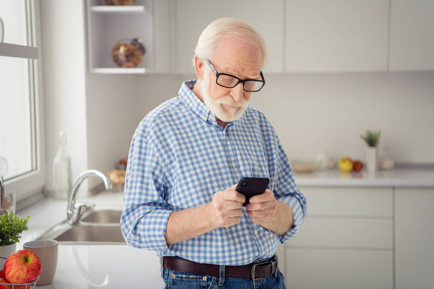 Close up portrait grey haired he his him grandpa concentrated hand arm telephone smart phone reader news observe wear specs casual checkered plaid shirt jeans denim outfit light room kitchen - Photo, Image