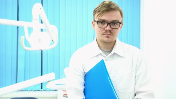 Portrait of a young male doctor standing in medical office and holding a blue folder under his arm, medicine concept. Dentist wearing glasses and white coat in his cabinet. - Footage, Video