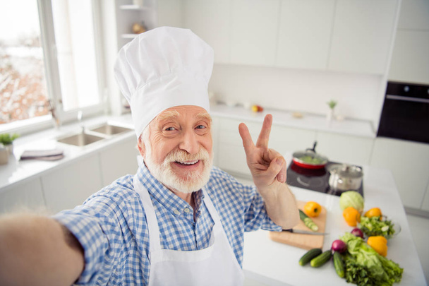 Close up photo cheer grey haired he his him grandpa telephone make take selfies video followers show v-sign say hi wear baker chefs costume casual checkered plaid shirt outfit house kitchen - Foto, Bild