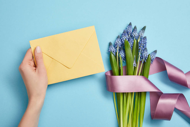 partial view of female hand with yellow envelope, and blue hyacinths with violet satin ribbon on blue  - Photo, Image