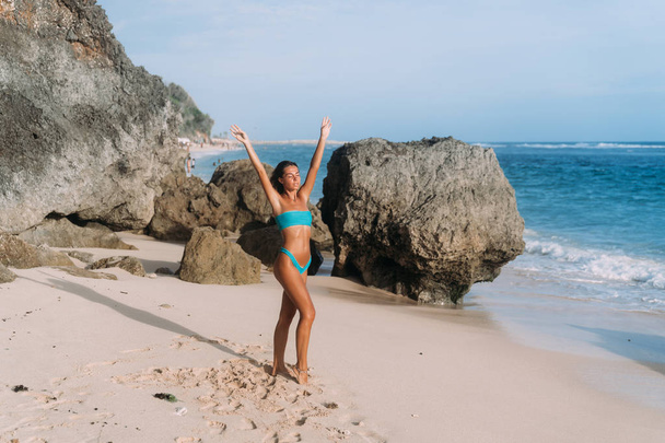 Slender tanned girl in blue swimsuit with raised arms standing on sandy beach with large stones - Foto, imagen