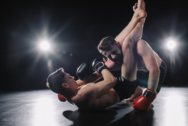 silná mma bojovník v Boxerské rukavice dělají bolestivé chokehold s nohama na jiný sportovec na podlaze - Fotografie, Obrázek