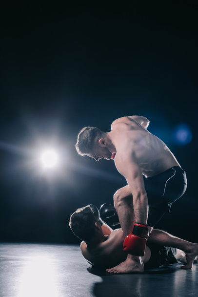 shirtless strong mma fighter in boxing gloves standing above opponent and punching him in head while sportsman lying on floor - Photo, Image