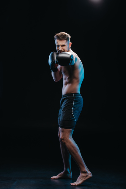muscular shirtless strenuous boxer in boxing gloves doing punch on black - Fotó, kép