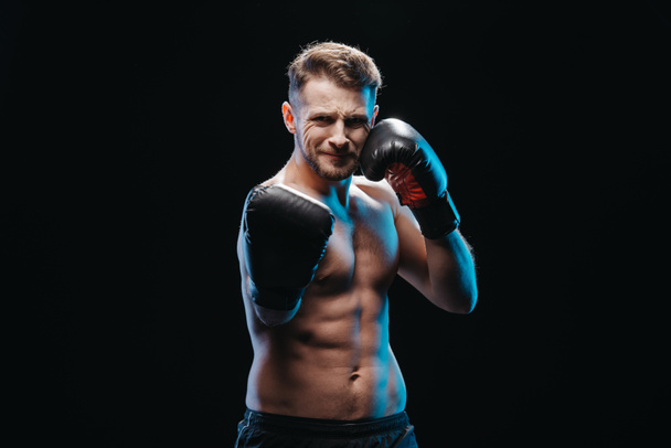 gespierde shirtless inspannende bokser in bokshandschoenen doen punch camera geïsoleerd op zwart - Foto, afbeelding