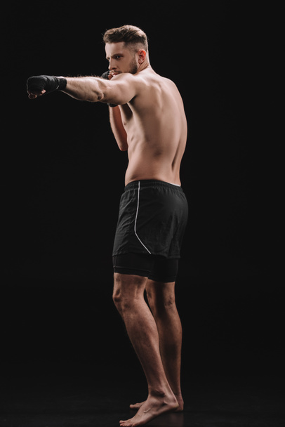 strong muscular mma fighter in bandages and shorts doing punch on black - Foto, Imagem