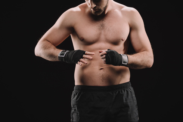 partial view of muscular sportsman looking at hands with bandages isolated on black - Photo, Image