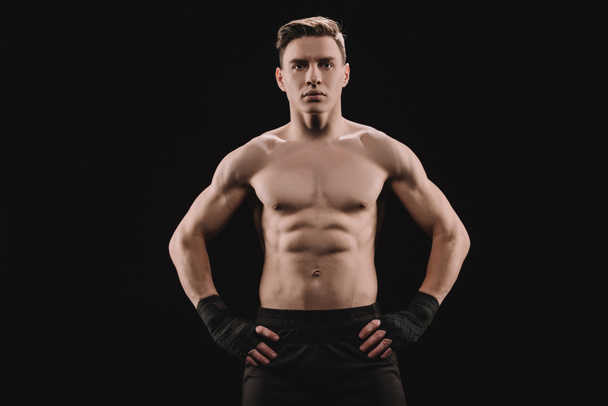 сильный мускулистый спортсмен, смотрящий в камеру с руками на бедрах, изолированных на черном
 - Фото, изображение
