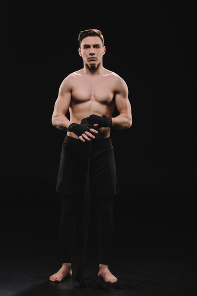 strong shirtless and barefoot muscular mma fighter fixing bandages and looking at camera on black - Photo, Image