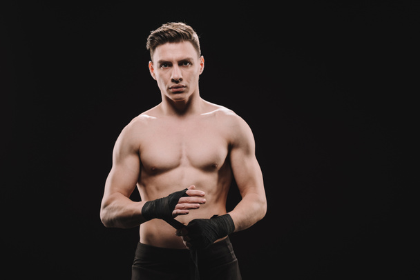 strong shirtless muscular mma fighter fixing bandages and looking at camera isolated on black - Photo, Image