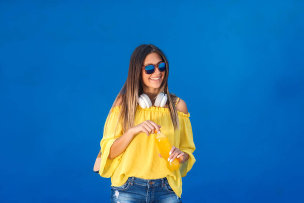 Porträt der schönen Brünette in gelber Bluse und mit Sonnenbrille und Kopfhörer um den Hals, die Saft vor blauem Hintergrund hält. - Foto, Bild