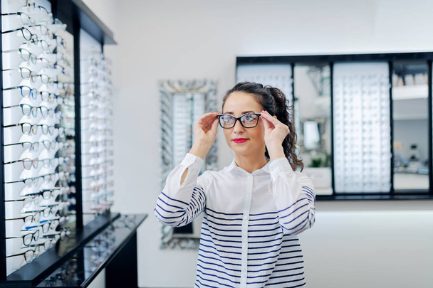 Portret van aantrekkelijke Kaukasische jonge vrouw brillen bij opticien winkel zetten. In achtergrond spiegel en brillen in rijen. - Foto, afbeelding