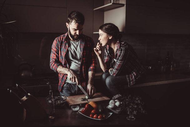 schöne junge Paar in der Küche zu Hause beim Kochen gesundes Essen. Der Mensch schneidet Salat. Frau sitzt auf Tisch und isst eine Gurke. Szene aus dem Familienleben. - Foto, Bild