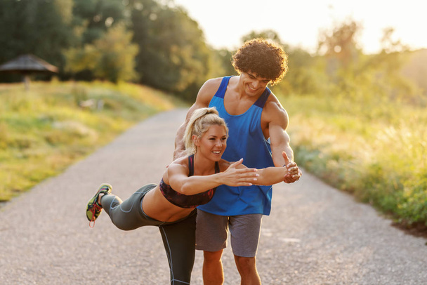 Man in blue t-shirt helping blonde woman with ponytail to do balance exercise in nature. Summer day. Three quarter length. - Photo, Image
