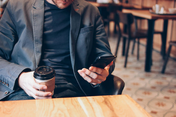 Erwachsener Mann trinkt Kaffee aus Pappbecher und benutzt Handy im Café - Foto, Bild