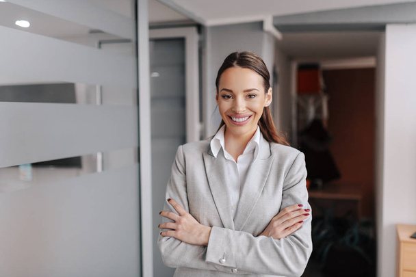 Charming Caucasian businesswoman in formal wear standing in office with arms crossed. You can't discover new oceans unless you have the courage to lose sight of the shore. - Foto, imagen