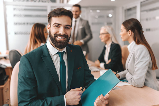 Smiling bearded Caucasian businessman in formal wear holding pen and folder while sitting in boardroom. In background colleagues talking. Wish for it and work for it. - Fotoğraf, Görsel