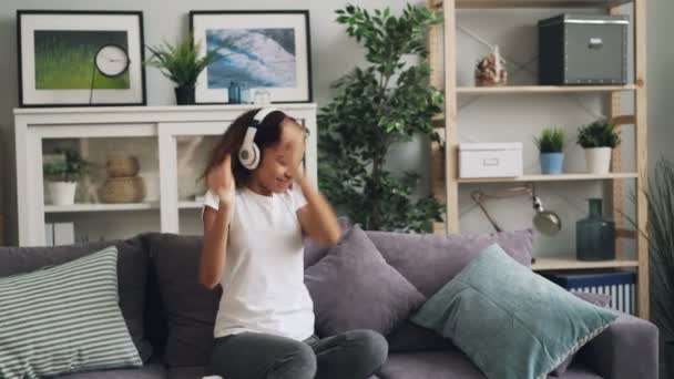 Happy African aAmerican lady in casual clothing is listening to music with headphones, dancing and relaxing sitting on couch at home. Modern technology and youth concept. - 映像、動画