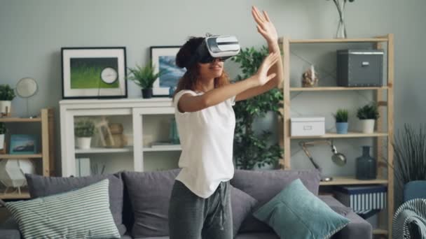 Young African American woman is enjoying experience with augmented reality glasses wearing modern vr headset. Girl is moving hands and head standing at home. - 映像、動画