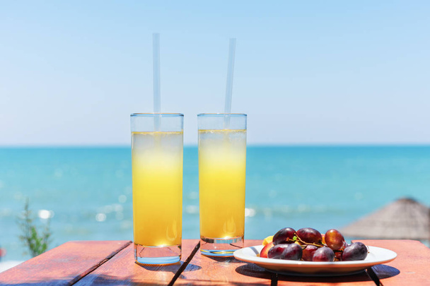 Two glasses of fresh tropical fruit juice on the beach, and a plate with grapes and watermelon on the table, against the sea. Holiday and vacation concept - Φωτογραφία, εικόνα