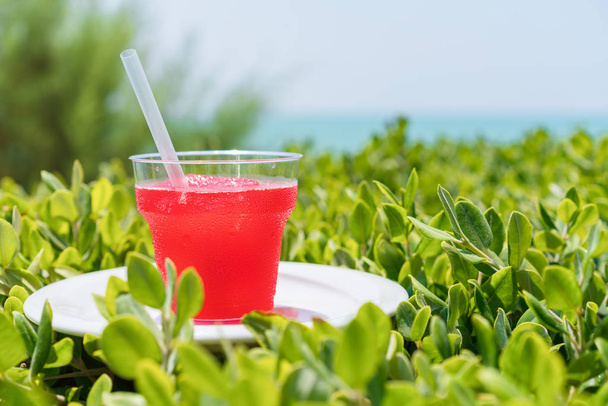 літо, склянка свіжого соку, з льодом, з червоних тропічних фруктів на пляжі, на зеленій траві, проти моря
 - Фото, зображення