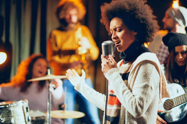 Mixed race woman singing. In background band playing instruments. Home studio interior. - 写真・画像